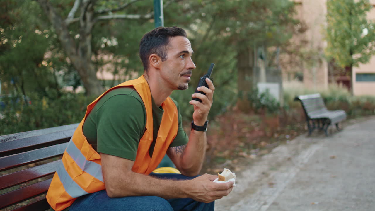 Relaxed contractor answering walkie-talkie in city closeup. Man holding sandwich