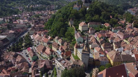 Beautiful-aerial-view-of-Sighisoara-old-town-in-Transylvania,-central-Romania-showing-colourful-buildings,-a-church-tower-and-a-hill-covered-in-trees