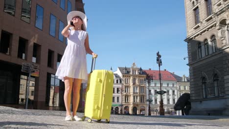 vertical video a slim young girl in a cute white dress and hat stands with a yellow suitcase in the old city. a french woman tourist takes a photo of the architecture of the old beautiful city