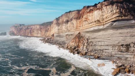 drone panning: crashing waves against rocky steep cliffs of nazare in portugal at golden hour