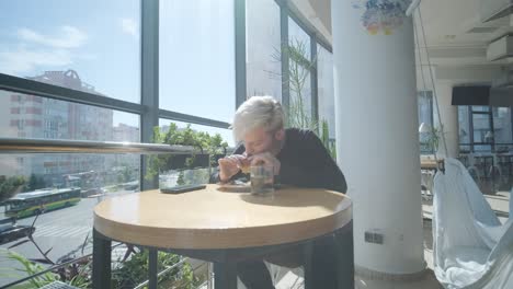 young man with blond hair enjoying a sandwich and iced coffee at a cafe table by a window with city views, natural light, and potted plants. relaxed and casual dining scene