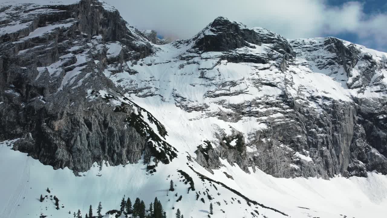Panorama flight along a glacier rock snow mountain top near Bavaria Elmau castle in the Bavarian Austrian alps on a cloudy and sunny day along trees and forest in nature with avalanches going down