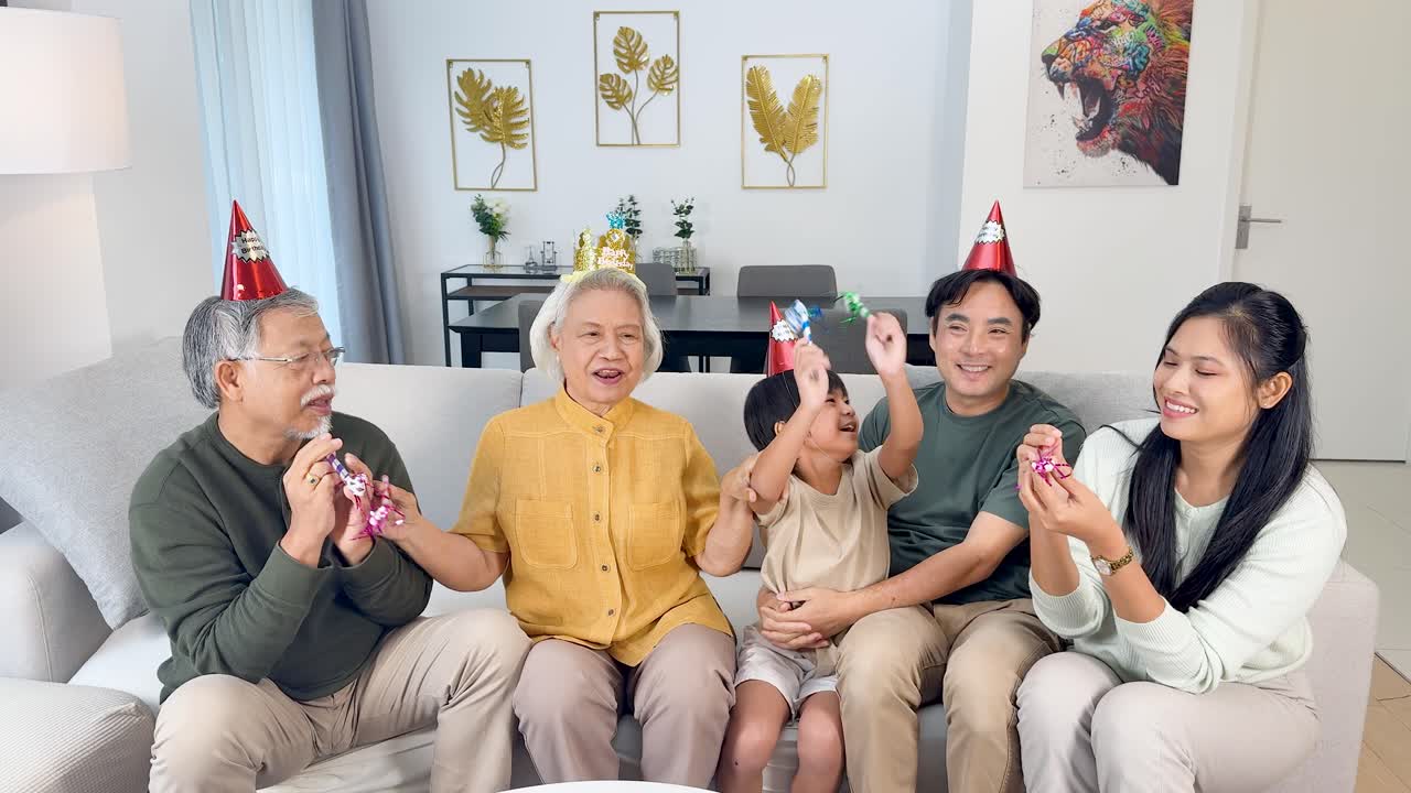 Four generations enjoy a joyful birthday party with hats, laughter, and party blowers indoors