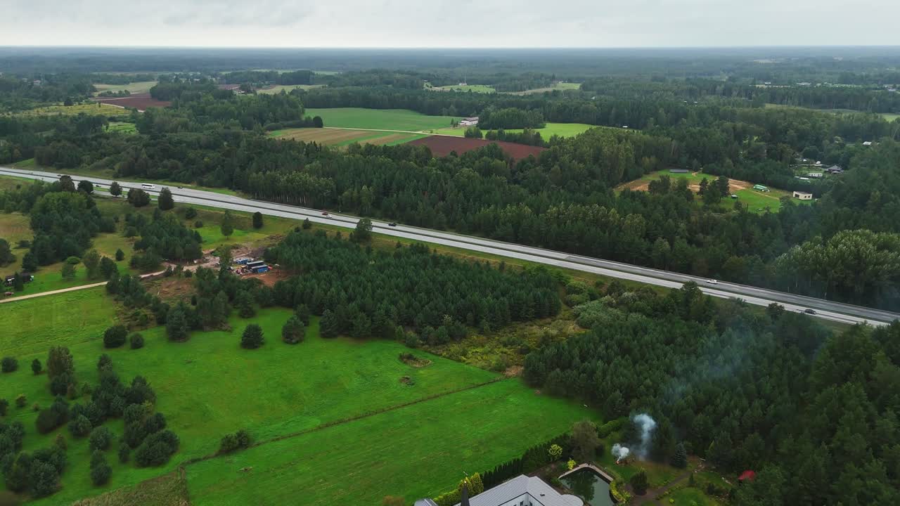 Ikskile’s town in Ogre Municipality, rural expanse and red building near the A6 highway surrounded by forest patches and farmland, blending urban infrastructure with natural textures, drone shot
