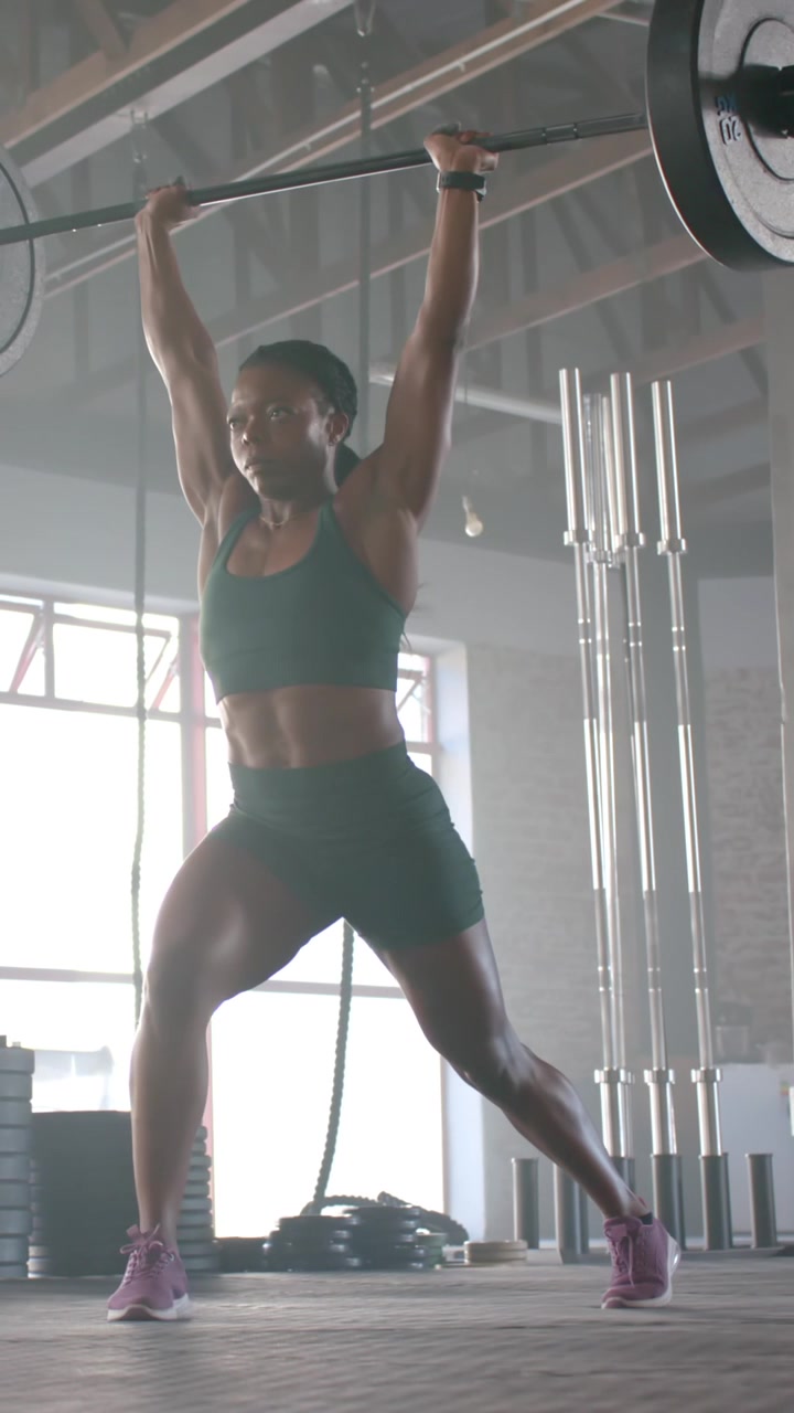 Video verticale: Donna afroamericana in abbigliamento da palestra che esegue uno slancio con bilanciere sopra la testa partendo dal front rack per l'equilibrio.