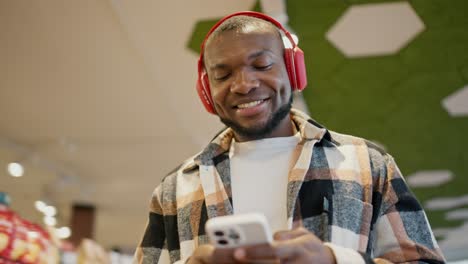 Shot-close-up-of-a-happy-man-with-Black-skin-color-with-a-beard-in-a-checkered-shirt-and-a-white-T-shirt-in-red-wireless-headphones-walks-through-a-large-supermarket-and-looks-at-his-grocery-list-on-a-white-phone