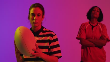 portrait of diverse female rugby players with rugby ball over neon pink lighting