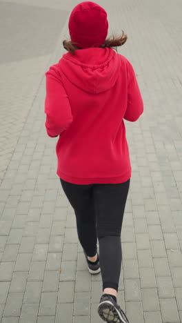 back view of woman jogging outdoors on interlocked pavement during winter, hair flowing as she runs, snow-covered ground, flower pot accentuating serene urban environment in cold weather