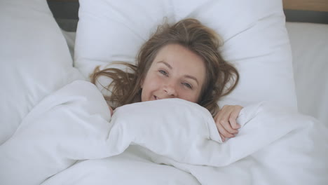 tanned smiling woman lying in bed under a blanket and peeks out from under him. pretty young woman peeking out from under the blanket smiling at camera.