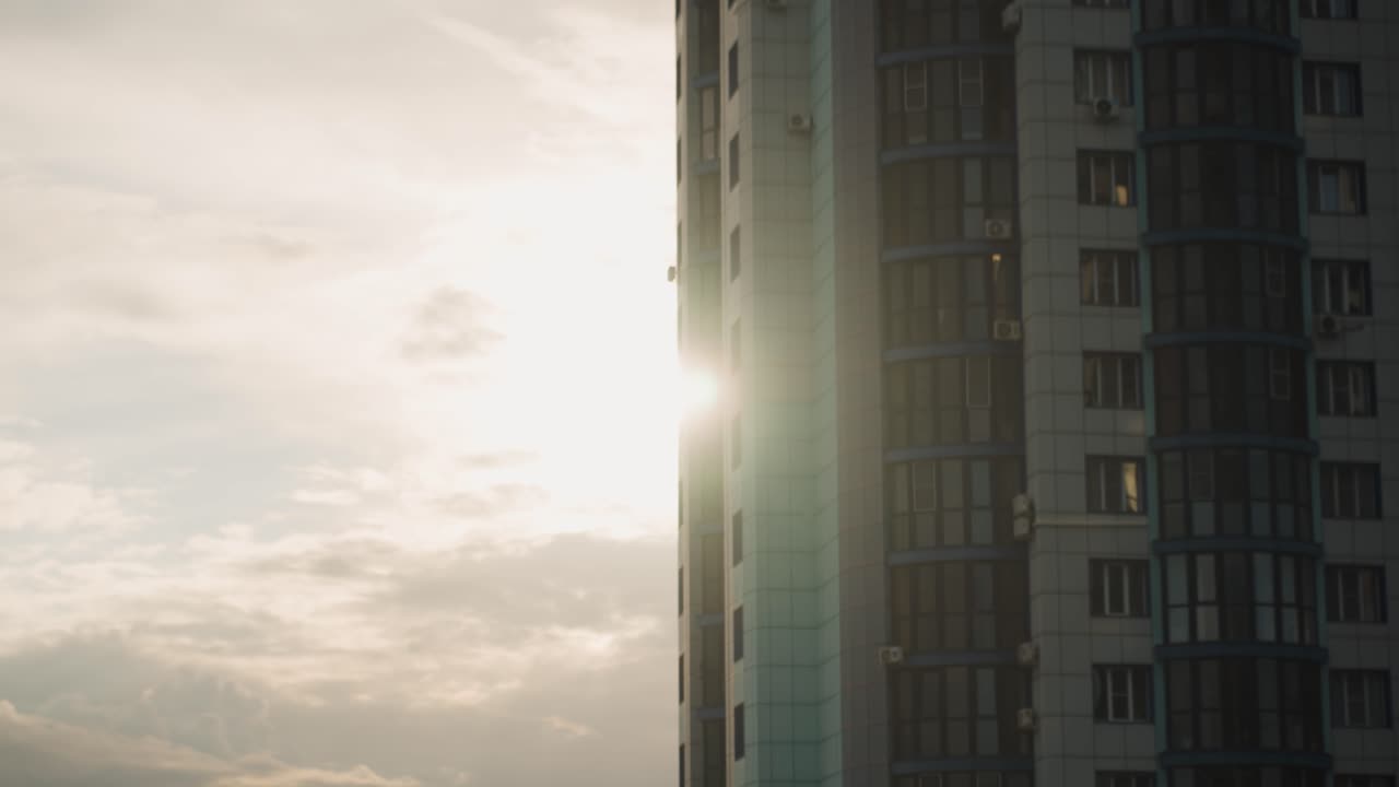 Luz do pôr do sol incidindo em torre residencial, com efeito flare. Brilho intenso através de uma fenda estreita entre fachadas, janelas refletoras capturando brilho quente. Nuvens carregadas se acumulando, silhueta de antena no telhado, ambiente tranquilo.