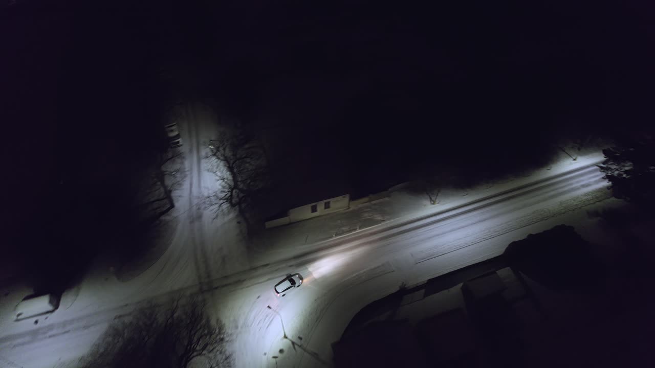 Drone follow shot following a white car on a dark night with the streets fully covered with snow on a cold wintry night.