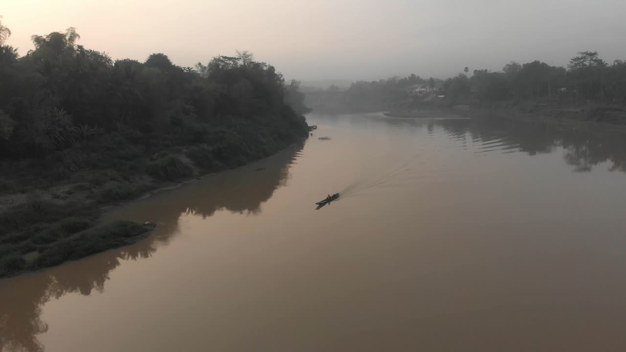 En liten lokal båt glir på en elv i Laos ved soloppgang, sett fra luften