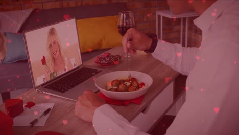 red heart icons floating over caucasian man having lunch while having a video call on laptop