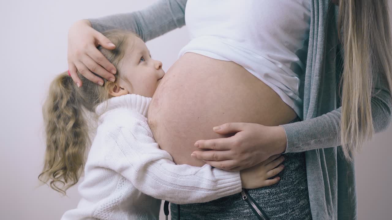 스웨터를 입은 소녀가 임신한 엄마를 안아서 집에서 배를 키스합니다.