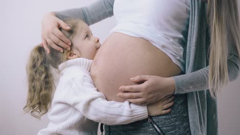 girl in sweater hugs pregnant mommy kissing belly at home