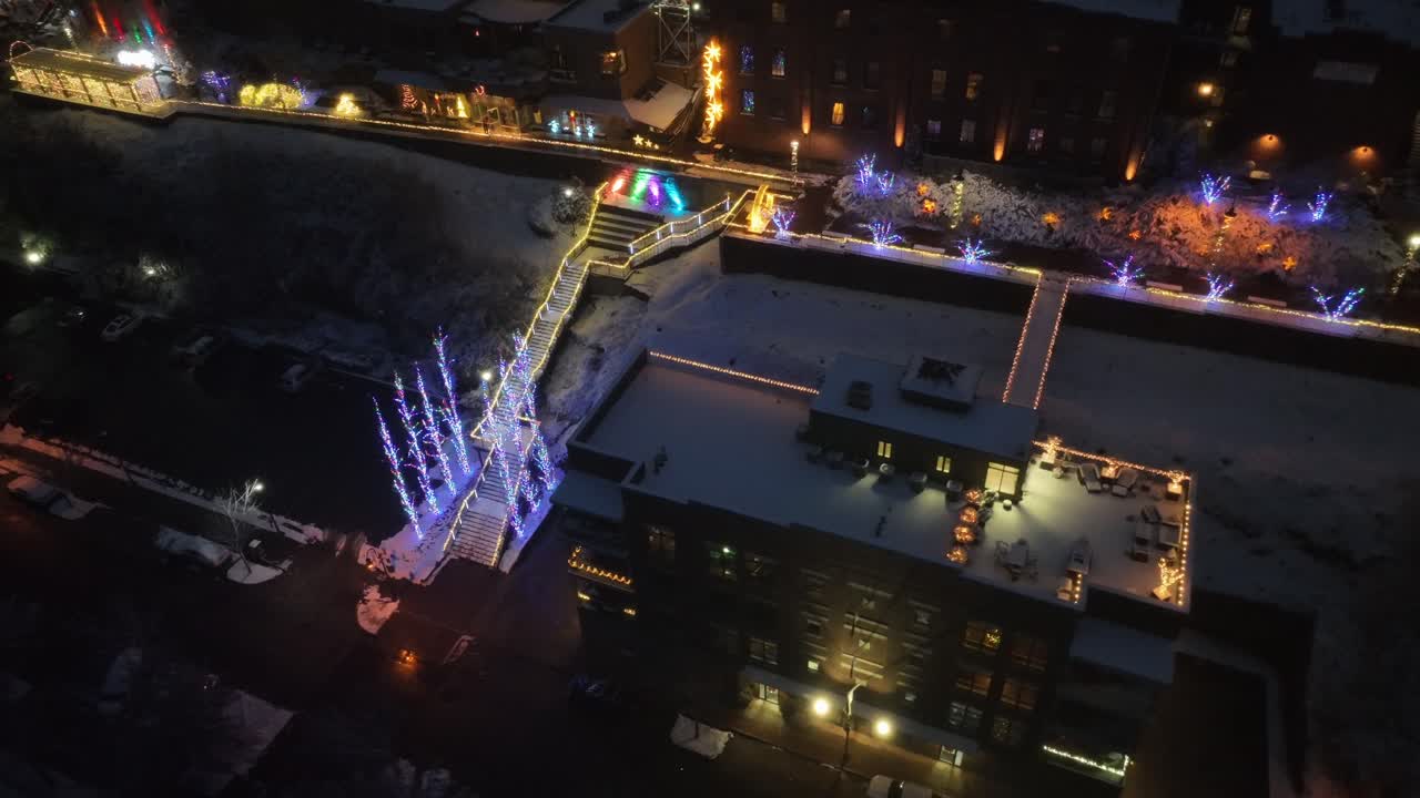 Snow-covered American city at night with glowing Christmas lights and illuminated walkways. Festive winter atmosphere in urban district during holiday season in United States. Wide shot. Aerial view