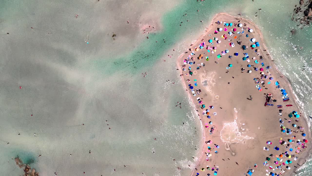 Groups of people relax and play on the sandy beach under the sun. Vibrant umbrellas dot the shore while many swim in the clear water, creating a lively atmosphere