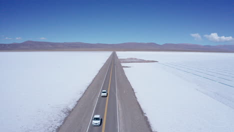 Cars-drive-on-straight-road-through-salt-flats-in-Argentina,-aerial