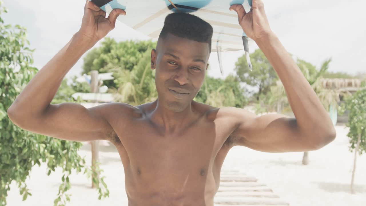 Ritratto di uomo afroamericano sorridente che trasporta una tavola da surf sulla testa in una spiaggia soleggiata, al rallentatore.