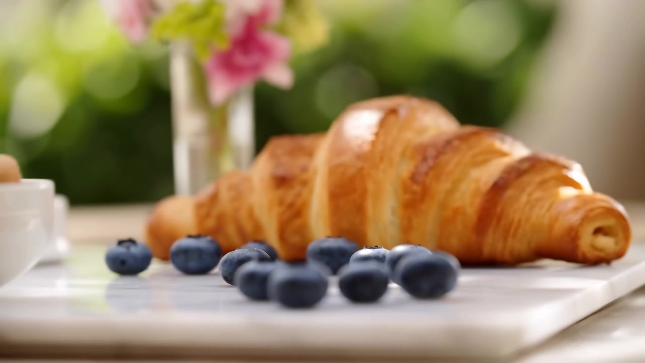 Mirtilli freschi riposano su un piatto bianco in primo piano, con un croissant, una tazza e dei fiori sfocati sullo sfondo sotto una luce naturale soffusa.
