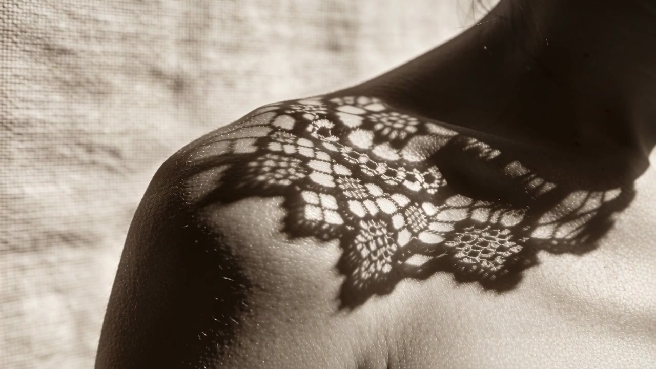 Encaje en movimiento proyectando sombra sobre el hombro y la clavícula de una mujer bajo una lámpara de estudio, creando un efecto artístico.