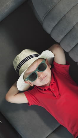 adorable child in summer straw hat relaxes on couch near suitcase in hotel room. cute little boy lies putting hands on occiput upper closeup