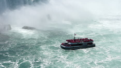 a bird flying over the maid of the mist cruise boat tour with tourists in red raincoats in basin of niagara falls, handheld shot