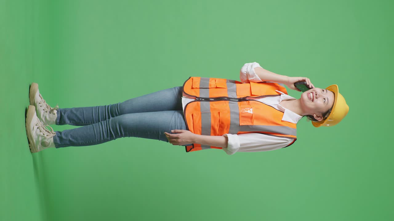vista lateral completa del cuerpo de una ingeniera asiática con casco de seguridad hablando en un teléfono inteligente mientras está de pie en el estudio de fondo de la pantalla verde