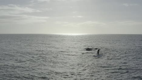 silueta stand up paddleboarder en un océano gris plano cerca del peligro de roca