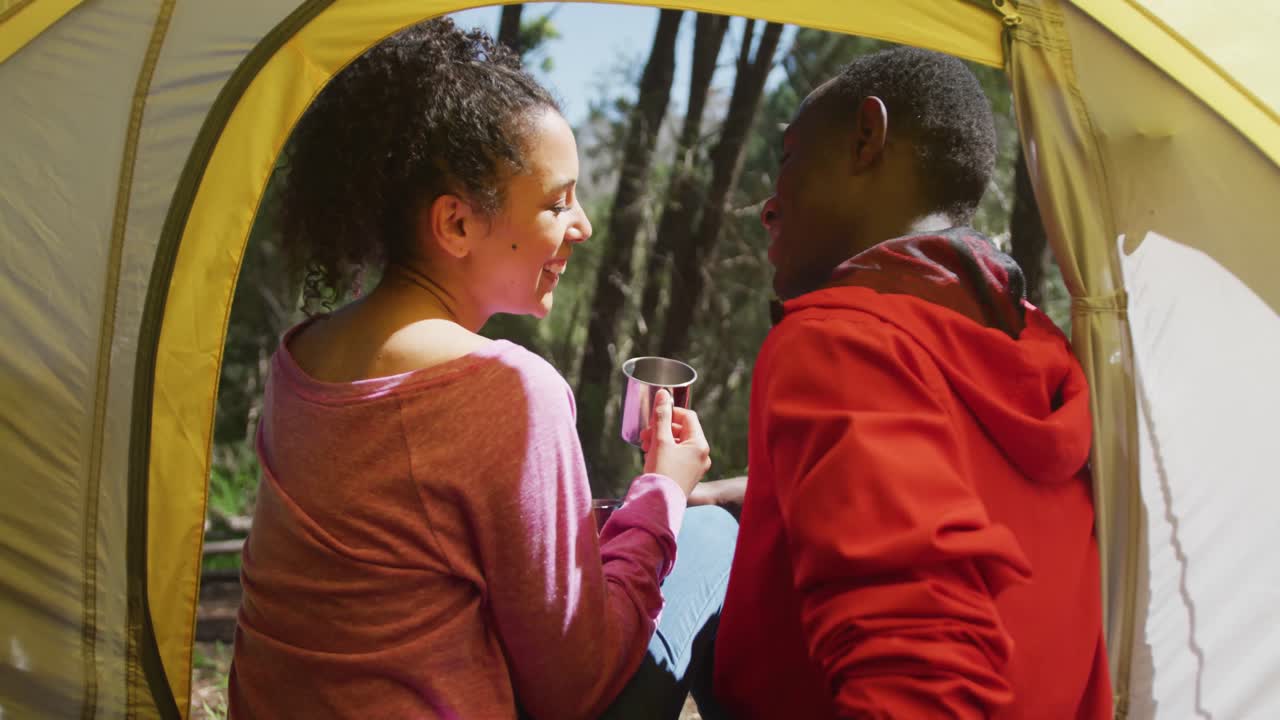 una pareja diversa sonriente sentada en una tienda de campaña y bebiendo té en el campo