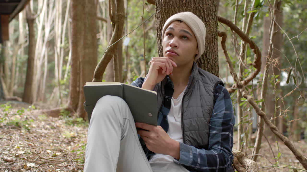 In forest, young man pondering ideas while writing in notebook under tree