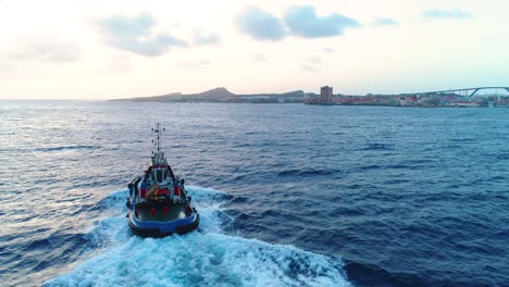 4k-Aerial-of-Tugboat-coming-into-Willemstad-city,-skyline-and-Queen-Juliana-bridge-in-background