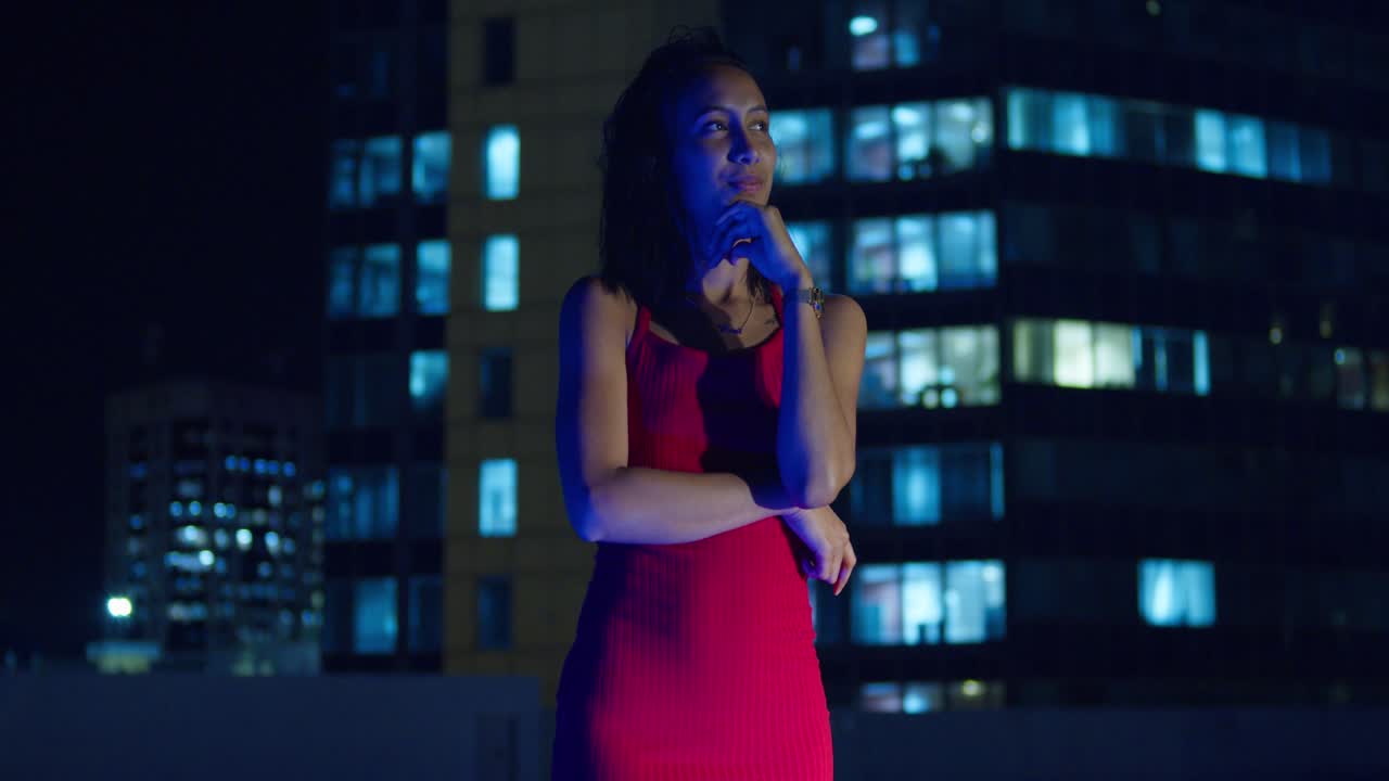 Su un tetto sotto il cielo notturno, una giovane ragazza con un vestito rosso si staglia contro gli edifici della città.