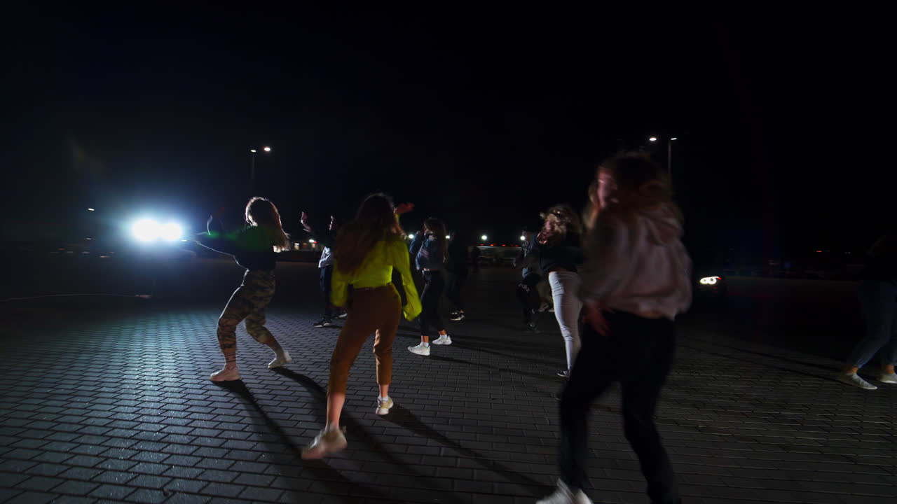 Large group of young people dancing hip hop on the square outdoors at night. Dance group performs in the flashing lights. Cars at backdrop.