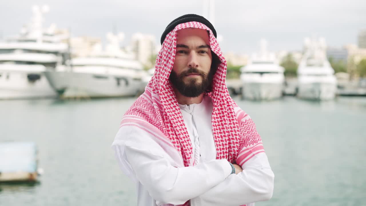 joven empresario saudí serio y confiado con ropa tradicional posando con los brazos cruzados en el puerto
