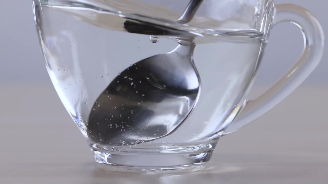 Primo piano di un cucchiaio di metallo che mescola lo zucchero nell'acqua calda all'interno di una tazza di vetro trasparente