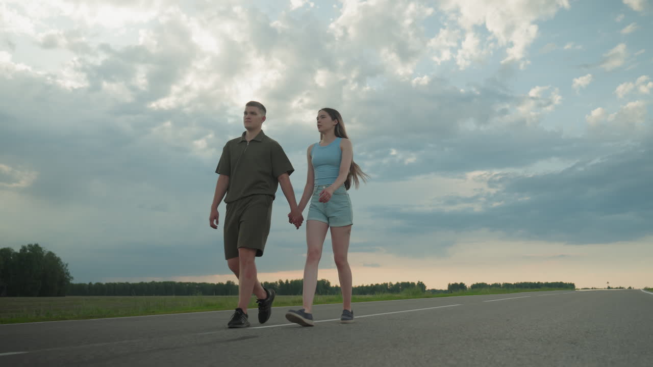 rear view of couple strolling empty country road hand in hand under dramatic sunset sky, lady glancing admiringly at partner in casual summer clothes