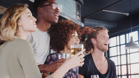 Grupo-De-Amigos-Y-Amigas-Celebrando-Mientras-Ven-El-Juego-En-La-Pantalla-En-El-Bar-Deportivo