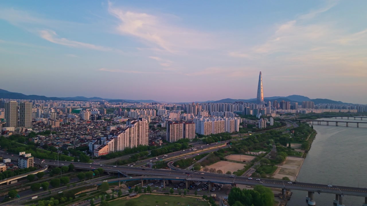 Ripresa aerea in parallasse che scivola sopra l'autostrada Olympic-daero e il Parco Gwangnaru Hangang al tramonto suggestivo, rivelando il vasto paesaggio urbano di Seul con la scintillante Lotte World Tower e il fiume Han