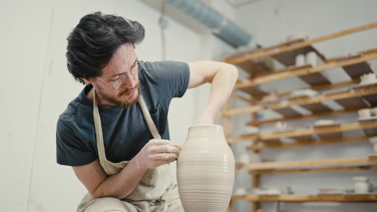 남자 마스터는 스튜디오에서 도자기 바에 세라 꽃병을 조각하고, 조각품을 완성하고, 느린 동작을 하고 있습니다.