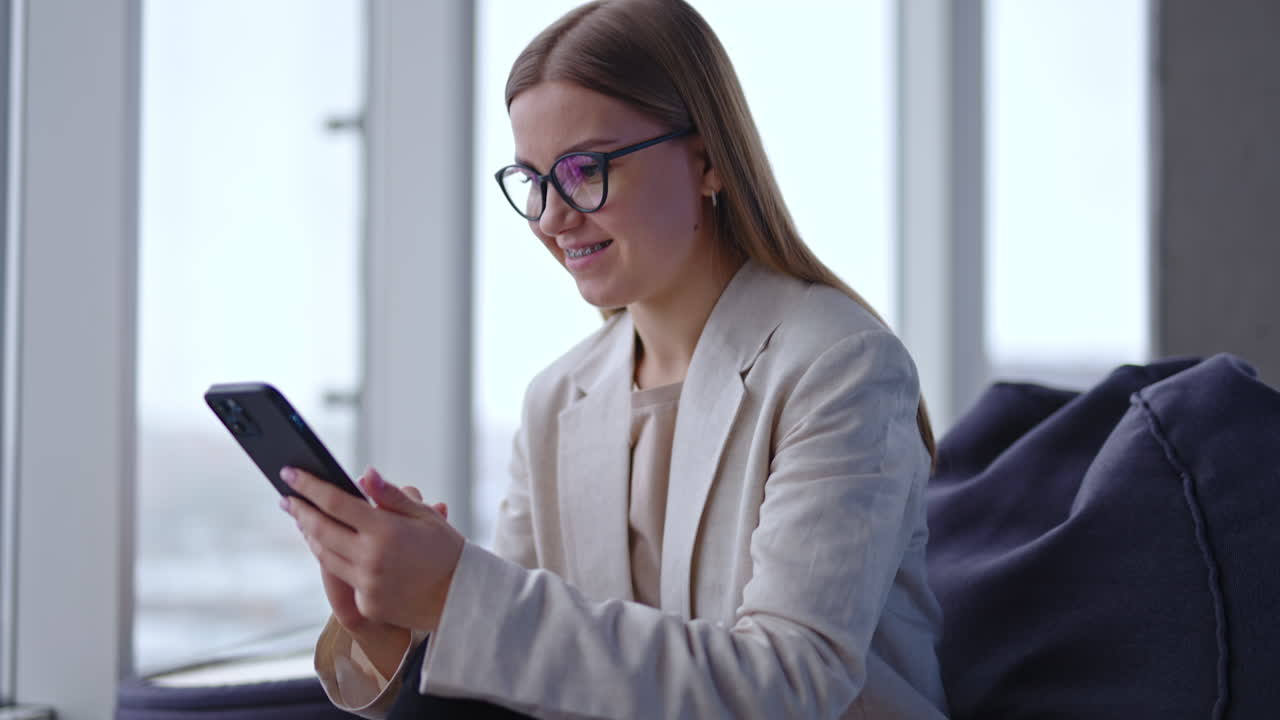 Beautiful young lady in glasses searches information on the phone. Woman looks at laptop and then opens her notebook. Light panoramic windows backdrop.