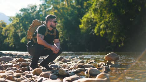 young hiker crouching by a river surounded by rocks and trees