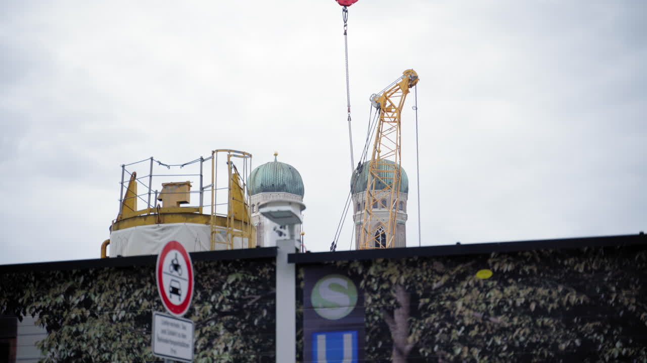 The iconic green domes of Frauenkirche in Munich rise above a construction site with cranes and scaffolding in the foreground. A contrast between historic architecture and modern urban development.