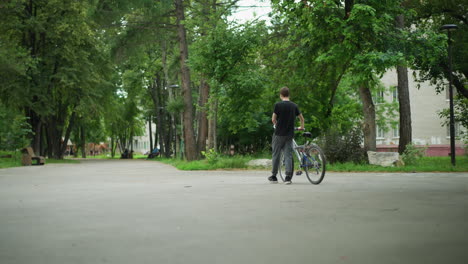 young boy in black top and ash pants is walking his bicycle along a park pathway, trees and greenery surround the scene, with blurred people in the distance, the background includes buildings