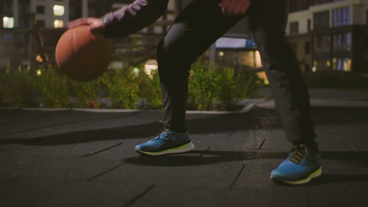 primer plano de un partido de baloncesto nocturno en la azotea, enfoque en la parte inferior del cuerpo, zapatillas azules y bote controlado sobre pavimento texturizado, luces de la ciudad difuminadas al fondo, ambiente tenue y melancólico, enfoque