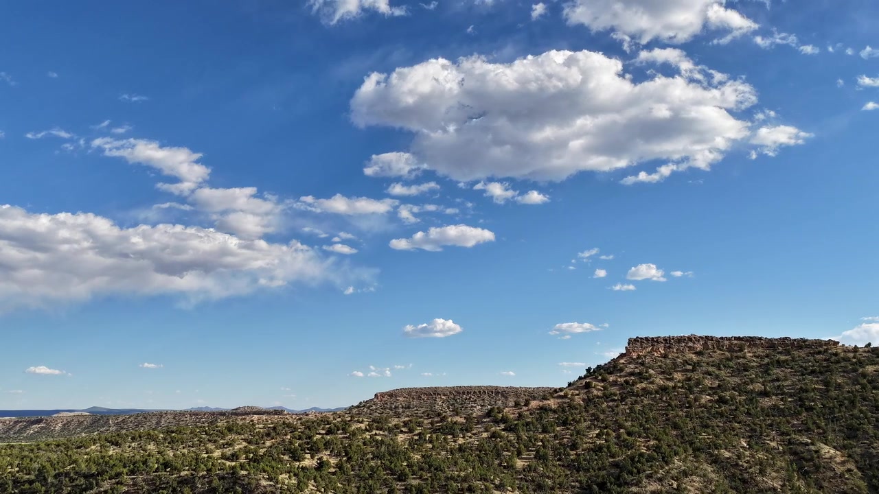 Un hyperlapse 4K con drone scivola in avanti per rivelare profonde mesas desertiche a strati sotto un vasto cielo blu con nuvole in movimento. Il movimento aereo cinematografico enfatizza la vasta scala del paesaggio aspro