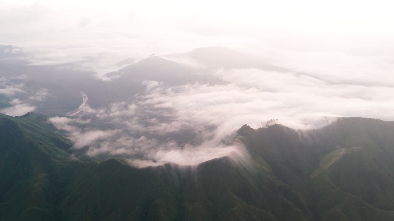 Breathtaking high-altitude aerial view of steep, rugged mountain ridges and peaks. Thick, swirling white clouds and fog roll dramatically through the deep valleys and crests of the mountains below
