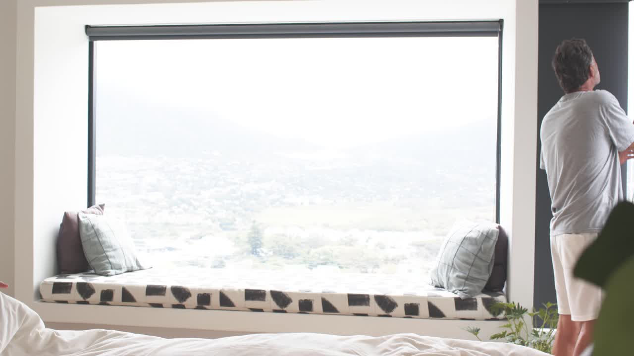 Morning light guiding woman making bed, glancing at window seat, man adjusting blind to view home