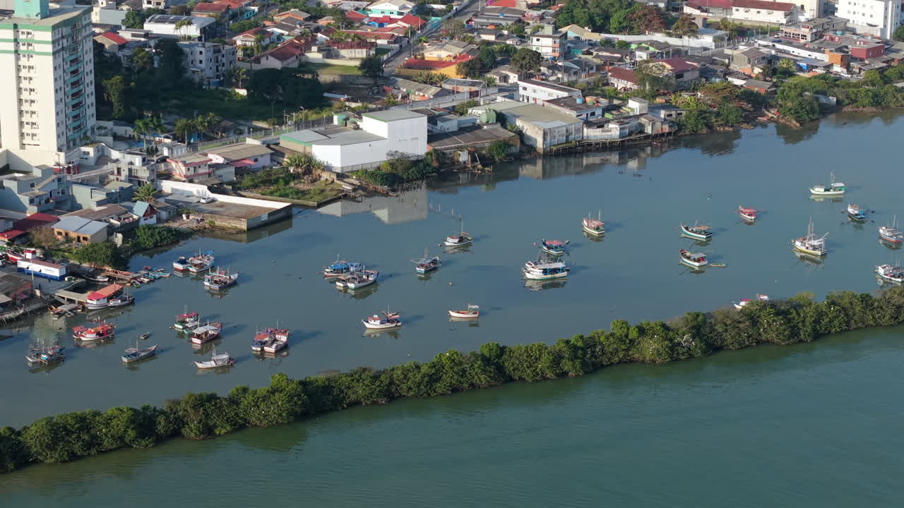 Aerial view of fishing boats resting on the calm waters of the Itajai river, surrounded by buildings and vegetation in Itajai, Brazil, during a sunny day