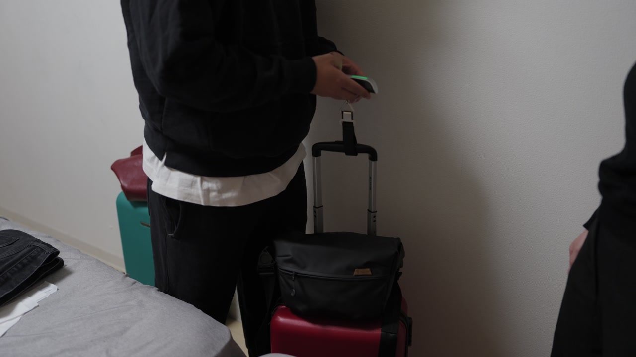Person using handheld luggage scale on stacked bags indoors, red and black suitcases visible